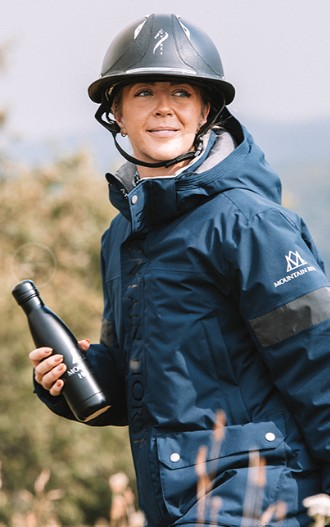 Ryttare i mörkblå Mountain Horse-jacka står utomhus med hjälm och vattenflaska, redo för ridning i alla väder.